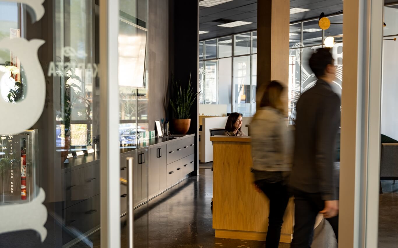 Wide view of a woman sitting at a front desk and smiling as two people walk past