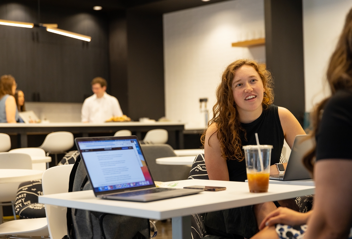 Two people sitting in a lounge area with their laptops and having a conversation while three coworkers converse in the background out of focus