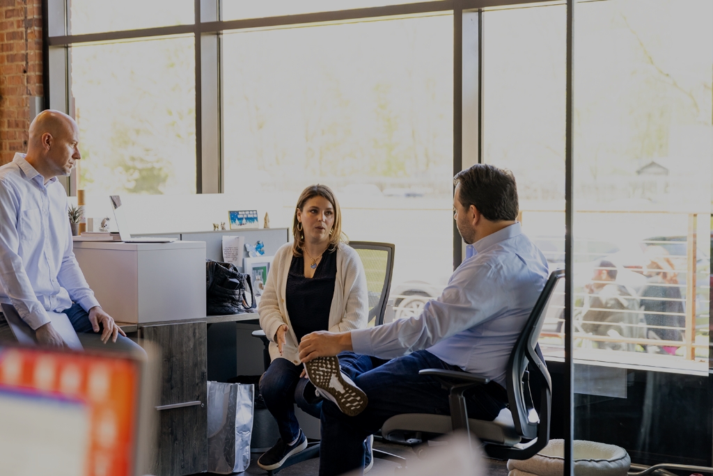 Wide view of three coworkers sitting in a group next to a desk and conversing